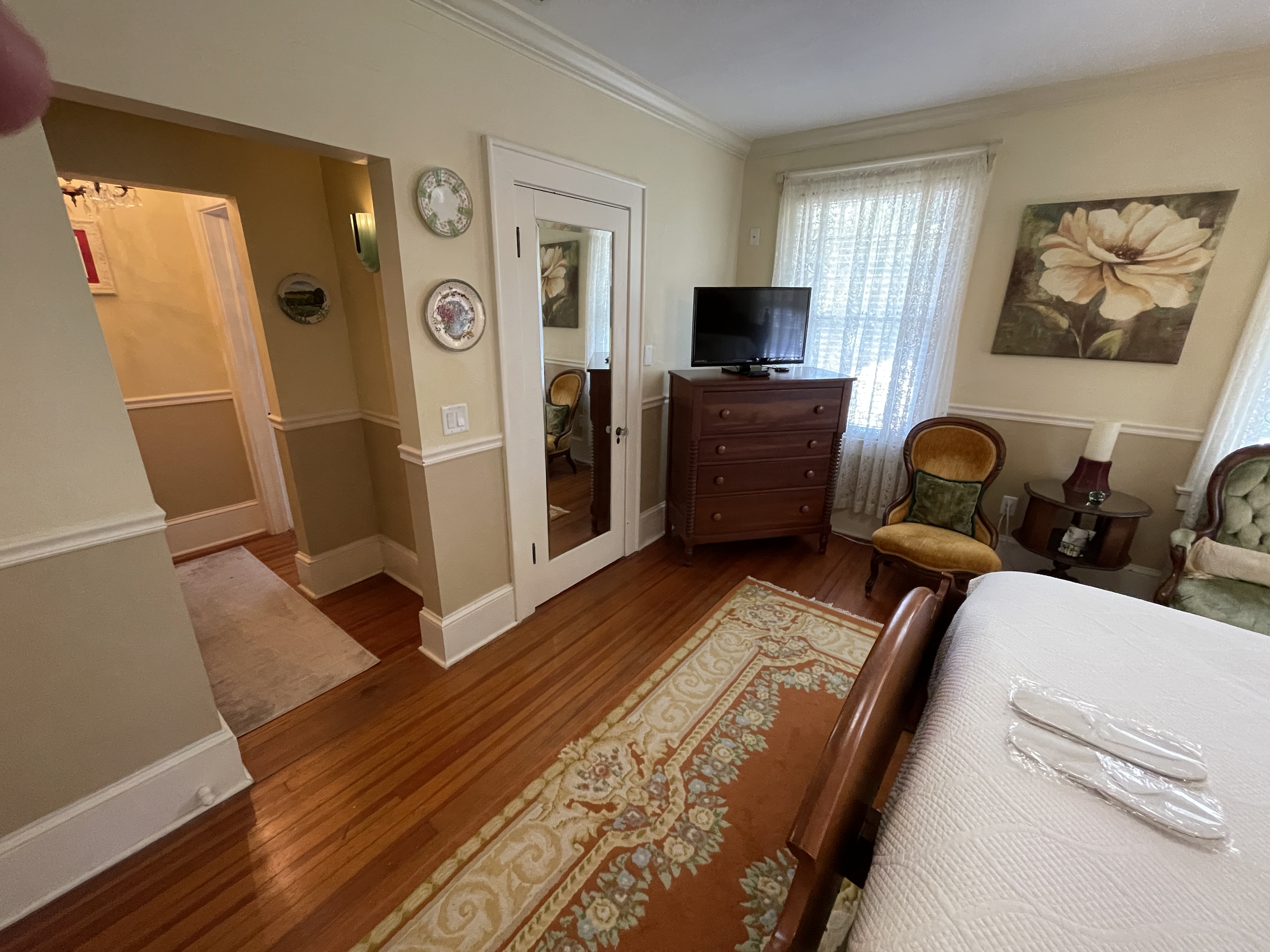 Cozy bedroom nook with classic wooden furniture, a TV, and an inviting rug. Leads to a bright hallway, offering a glimpse of charming inn living.