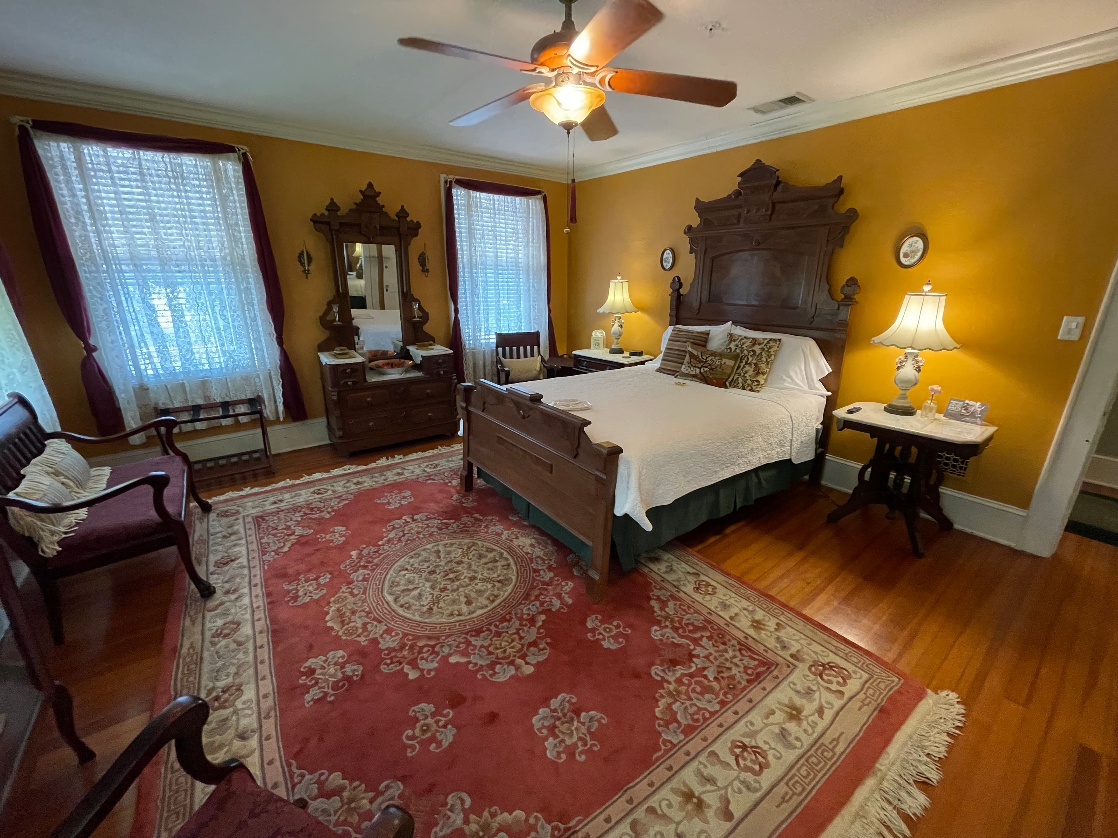 Elegant, antique-styled bedroom featuring a grand wooden bed, ornate headboard, classic wooden furniture, and a rich red rug.