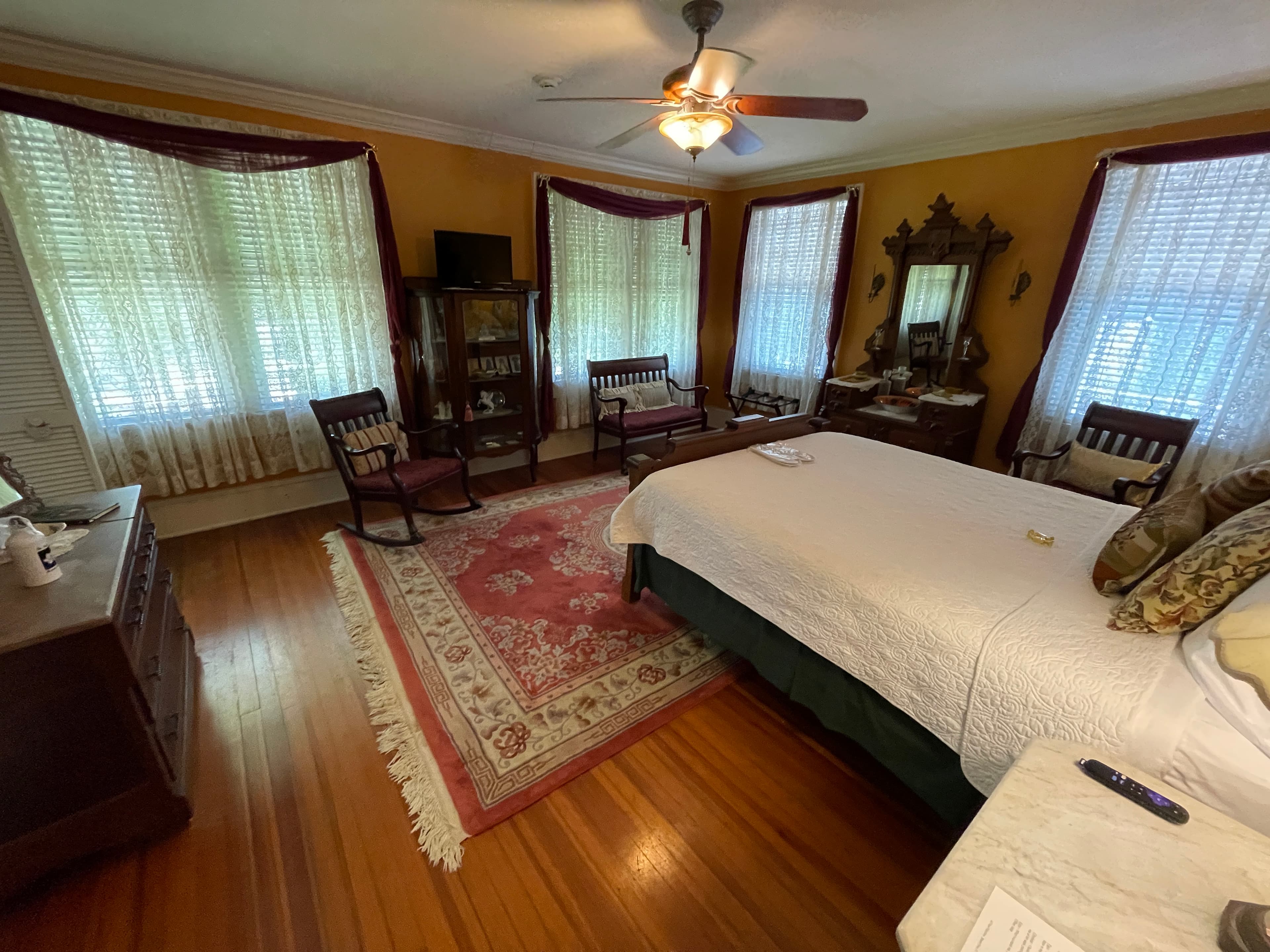 Spacious bedroom with a large bed, antique wooden furniture, and a vibrant patterned rug. Warm golden walls and ample natural light create a cozy retreat.