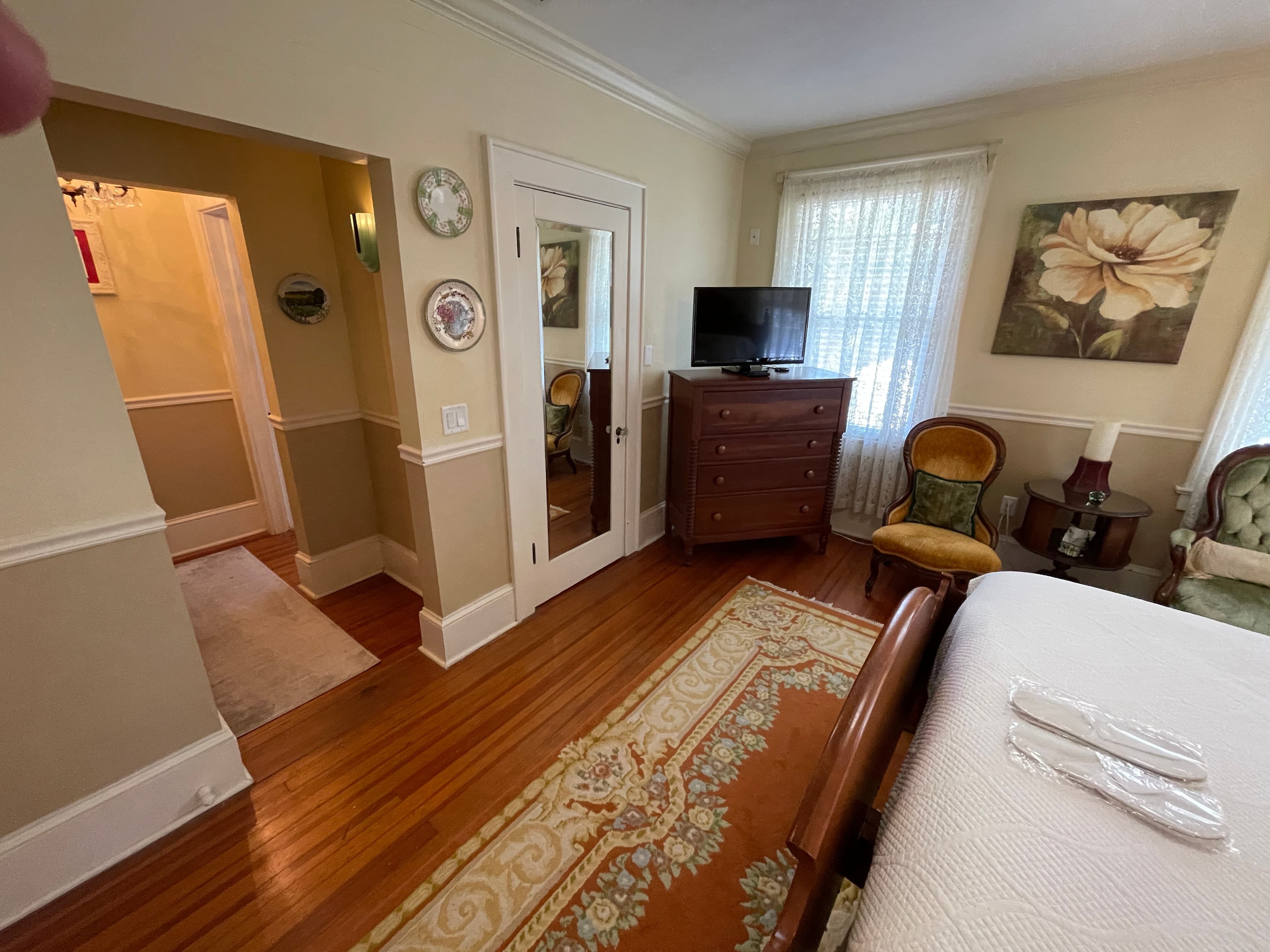 Cozy bedroom nook with classic wooden furniture, a TV, and an inviting rug. Leads to a bright hallway, offering a glimpse of charming inn living.
