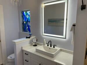 Contemporary bathroom vanity with a sleek vessel sink, backlit mirror, and modern black fixtures. A unique blue art window adds a pop of color to the bright space.
