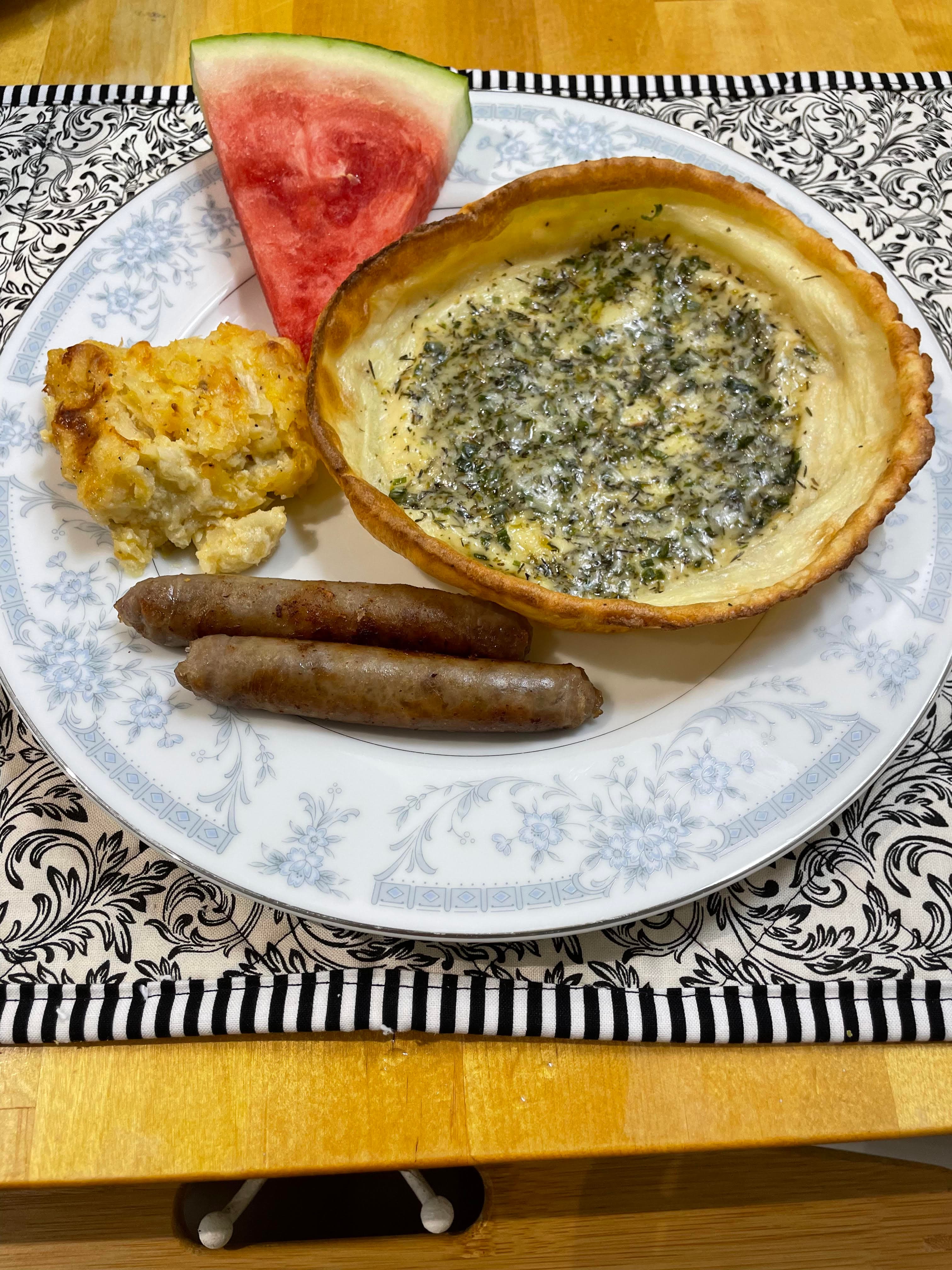 A plate with a slice of watermelon, two sausage links, a serving of cornbread, and a round quiche-like dish on a patterned tablecloth.