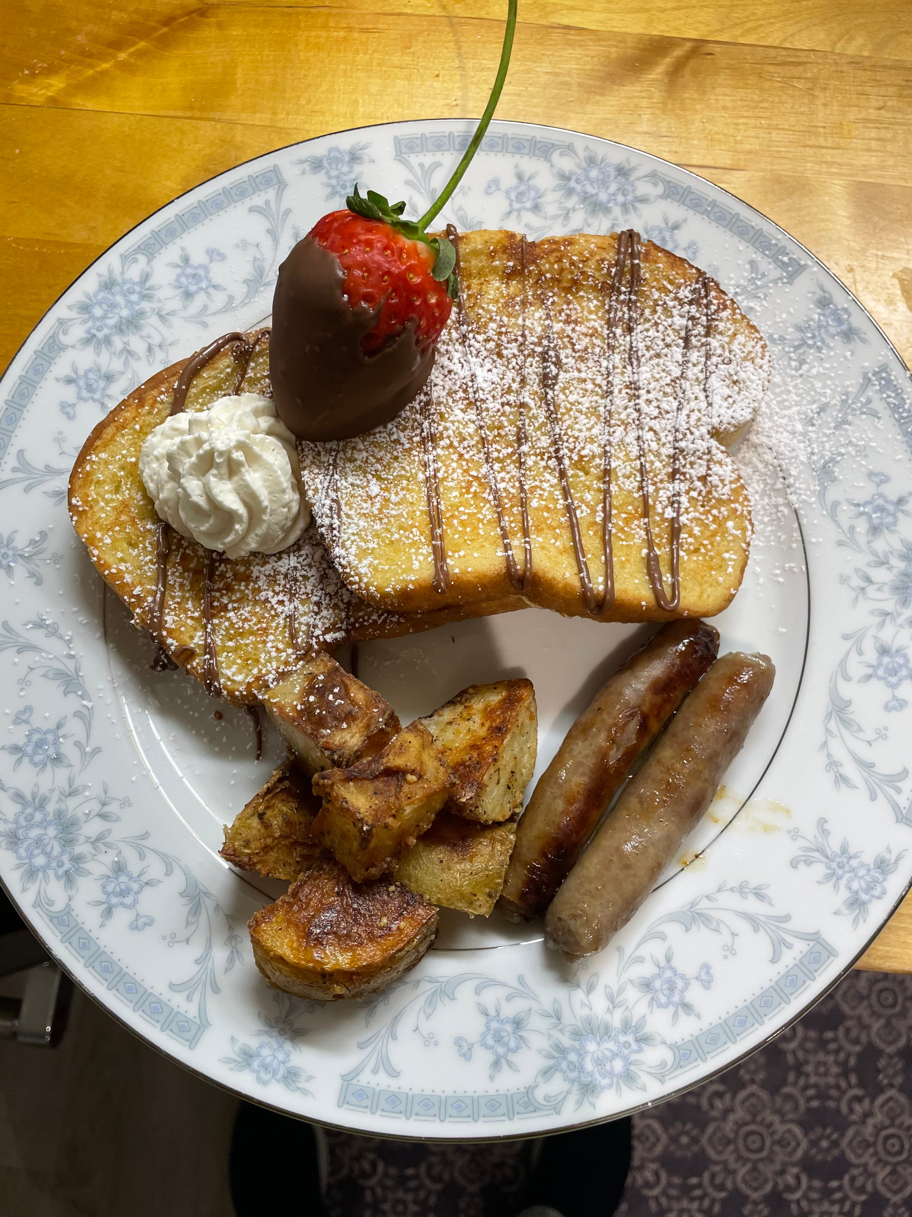 A decorative plate with French toast topped with a chocolate-covered strawberry, whipped cream, and powdered sugar, alongside sliced roasted potatoes and sausage links.