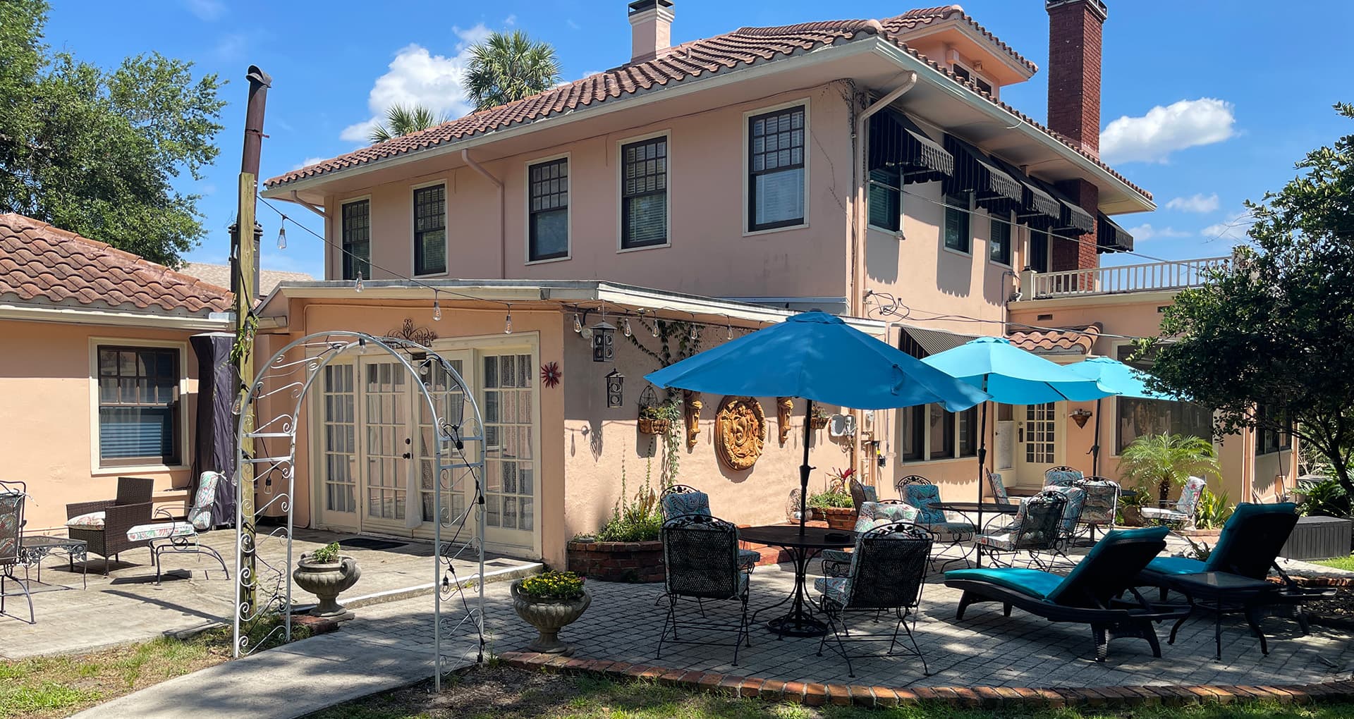 A pink two-story house with a patio featuring blue umbrellas, seating areas, and an archway.