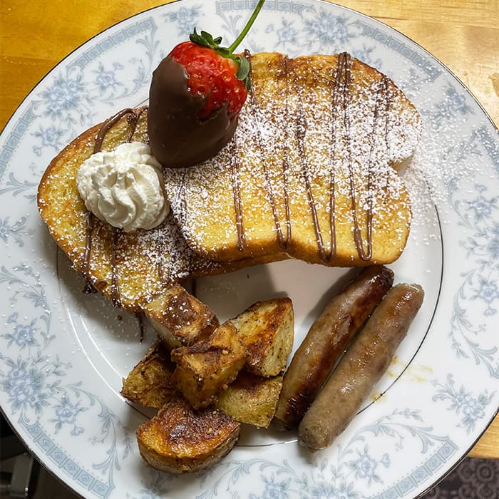 A plate of French toast topped with a chocolate-covered strawberry, whipped cream, golden potatoes, and sausages.
