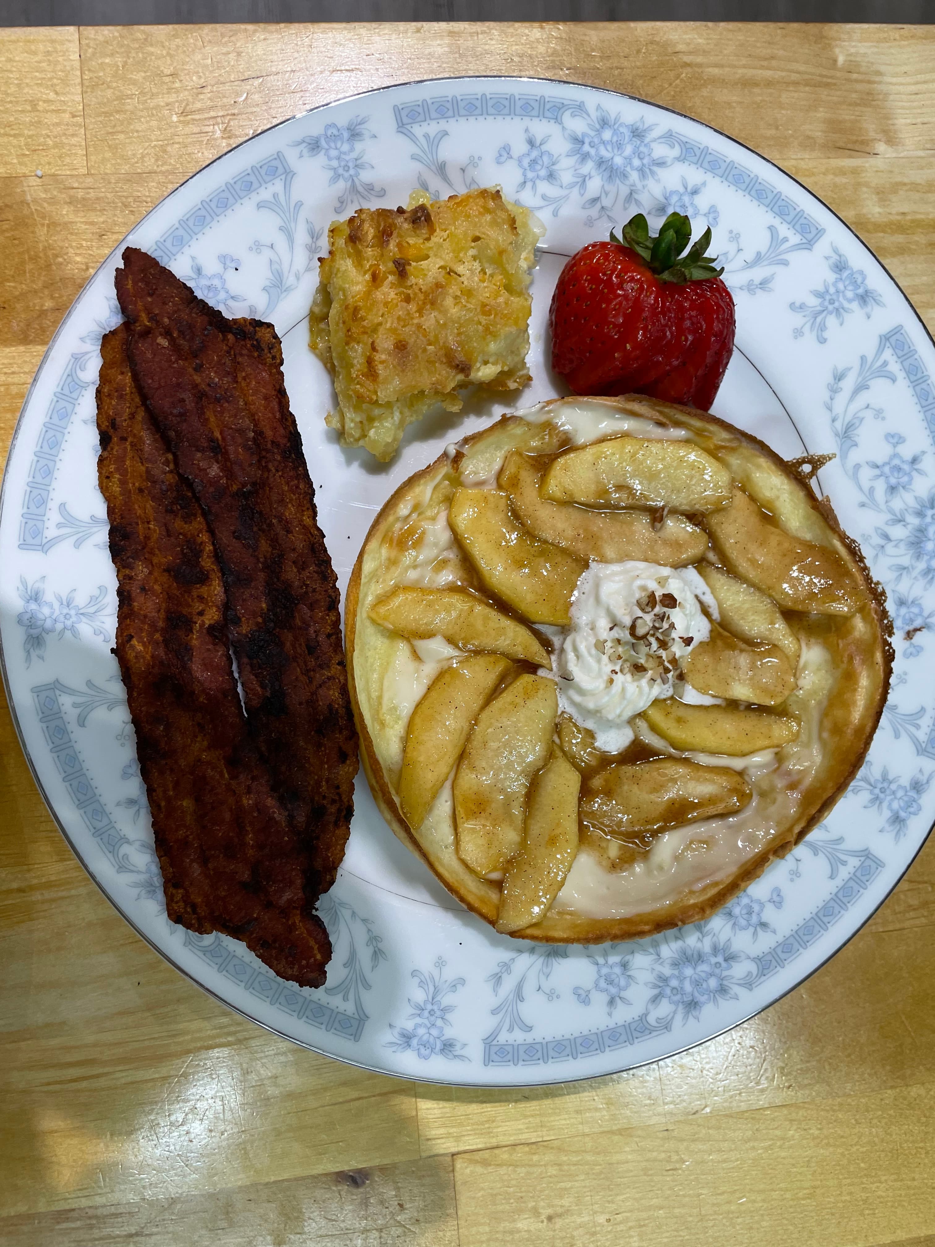 A plate featuring pancakes topped with apple slices and whipped cream, crispy bacon, a slice of casserole, and a fresh strawberry.