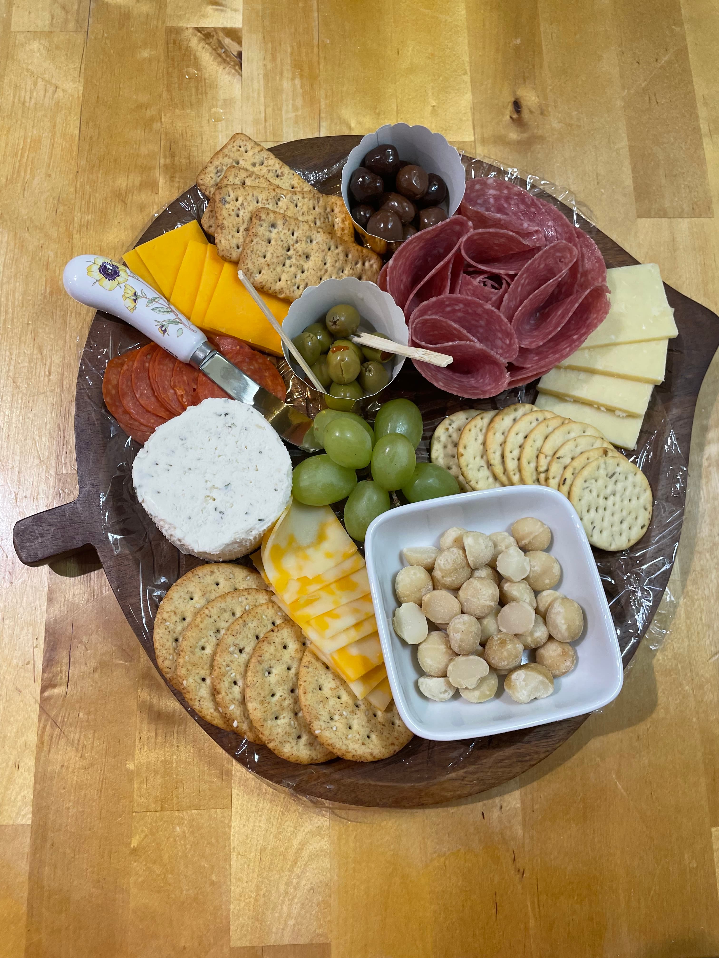 A wooden platter featuring an assortment of cheeses, crackers, meats, olives, grapes, and macadamia nuts.