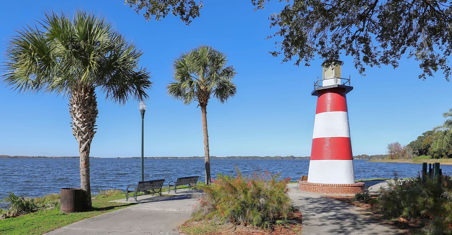 A red and white lighthouse stands by the water, surrounded by palm trees and benches under a blue sky.