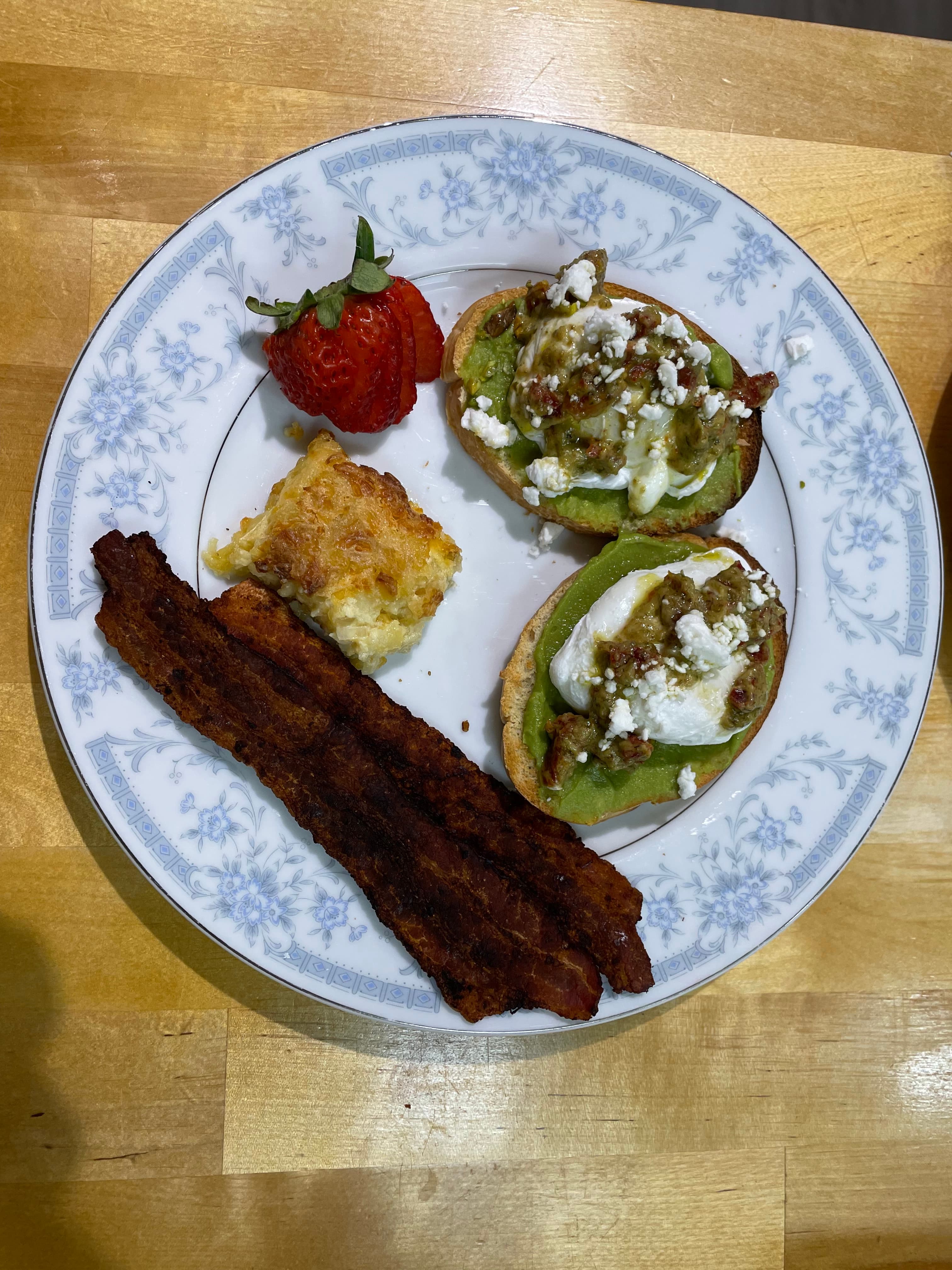 A plate featuring two avocado toast halves topped with poached eggs and feta, crispy bacon, a slice of baked dish, and a strawberry.