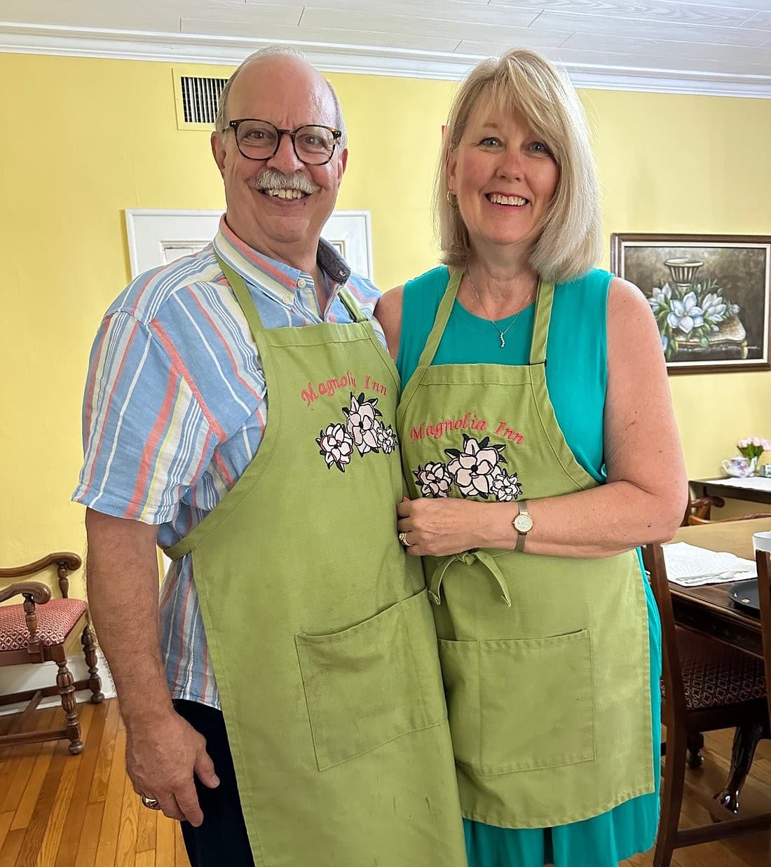 A smiling couple poses together in green aprons featuring floral designs, standing in a brightly lit room.