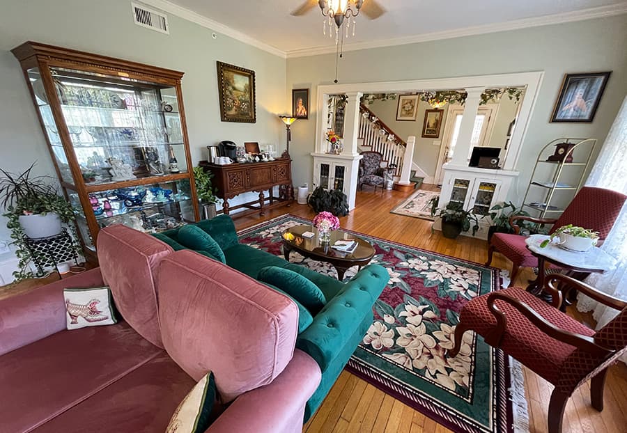 A cozy living room featuring green and pink sofas, a decorative rug, and a display cabinet, connecting to a hallway with a staircase.