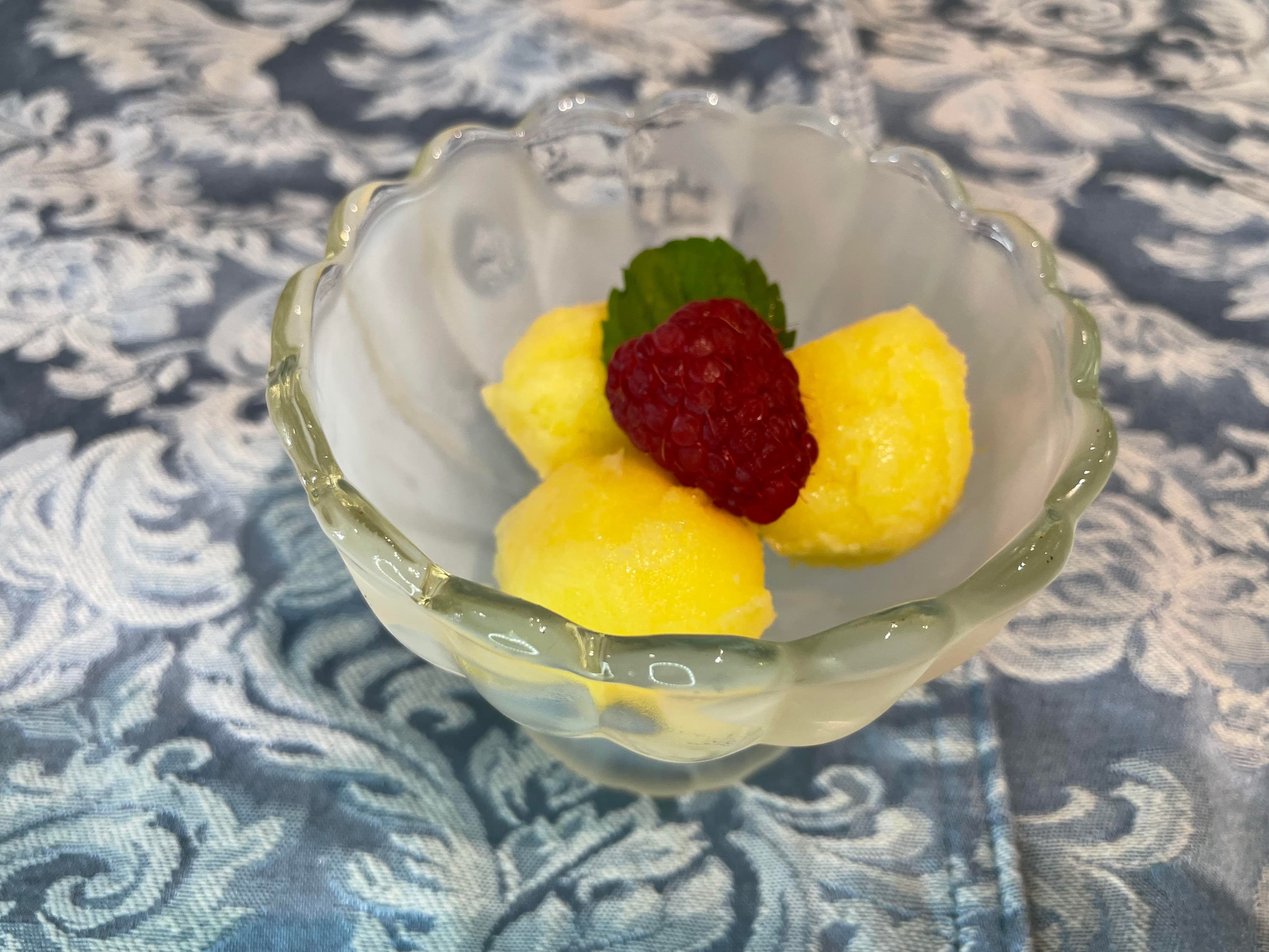 A glass bowl contains three scoops of yellow sorbet garnished with a raspberry and a mint leaf.